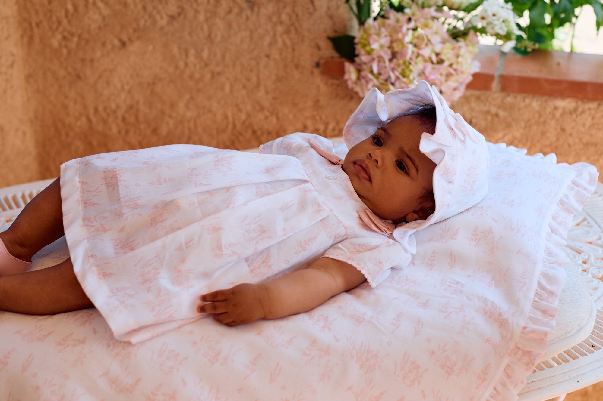 Baby Girl laying on a pink and white toile blanket in a matching dress and bonnet
