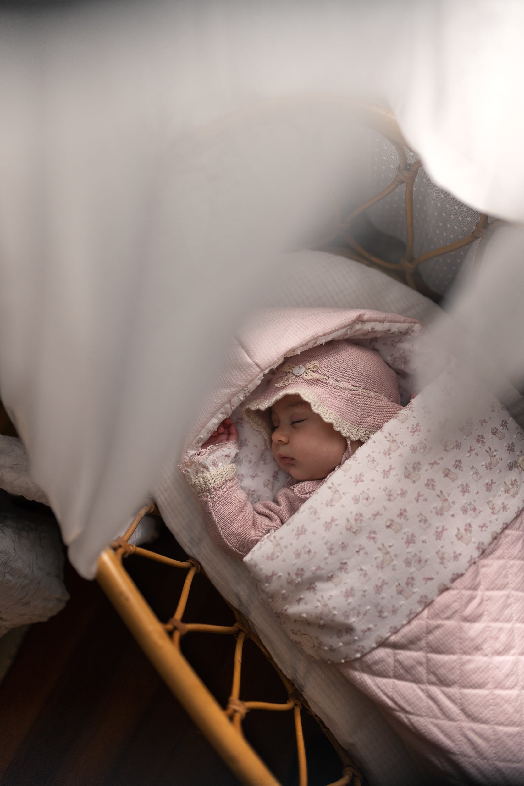 A sleeping newborn baby wrapped in a light pink floral blanket and wearing a matching pink bonnet, lying in a wooden bassinet under soft white drapery.