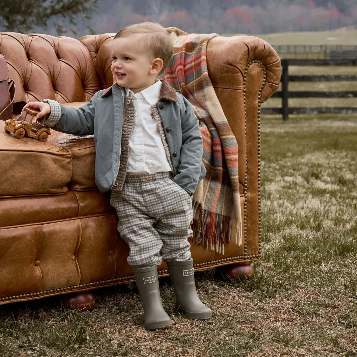 Child in formal attire standing next to a brown leather sofa outdoors.