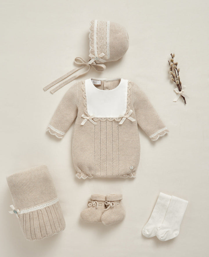 A coordinated baby clothing set laid flat on a warm cream background, including a beige knit romper with a white bib panel, a matching bonnet with lace trim and ribbon tie, a knitted bootie, a pair of white socks, and a folded knit blanket or swaddle. A small dried floral stem decorates the right side.