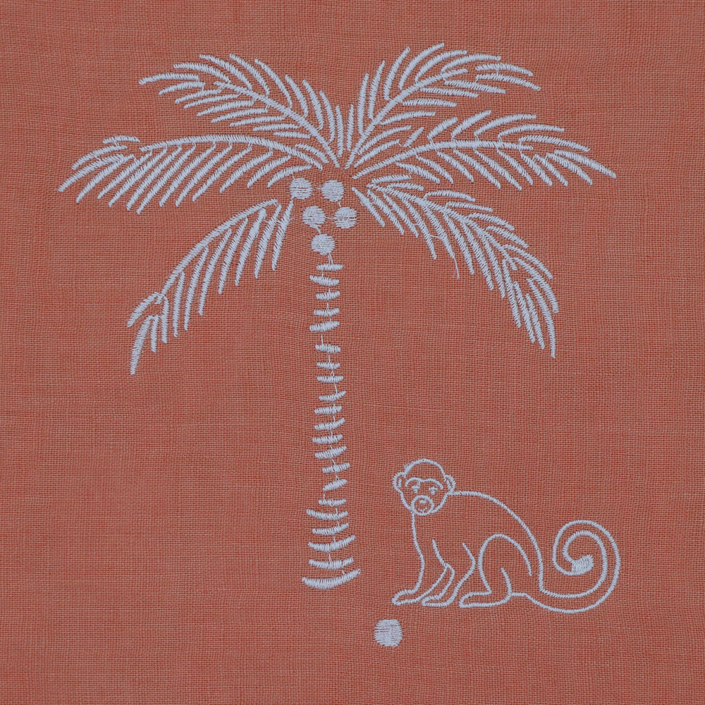 A close-up of a terracotta-colored woven fabric featuring a white embroidered palm tree with coconuts and a white embroidered monkey.