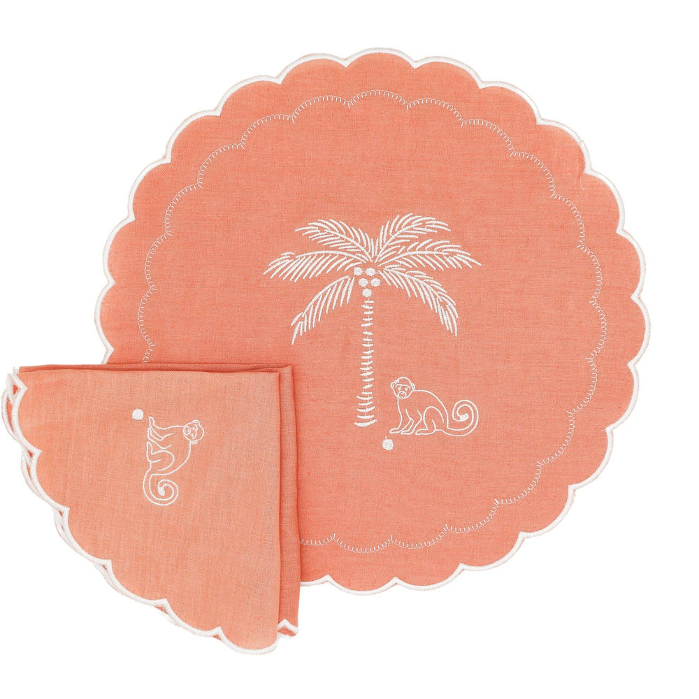 A coral pink linen table setting featuring a large round placemat and a matching folded napkin, both decorated with white embroidered palm trees, coconuts, and a monkey.