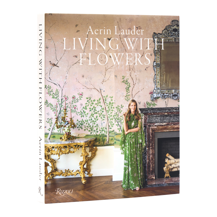 Acrin Lauder's 'Living with Flowers' book, published by Rizzoli, shown standing upright. The cover features a woman in a green floral dress standing in front of a fireplace with a large floral wall mural in the background.
