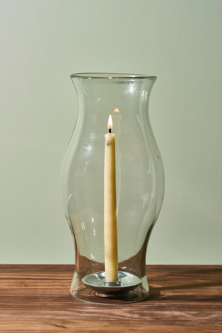 A clear glass candle holder vase with a lit cream taper candle inside, set on a wooden surface against a light sage green background.
