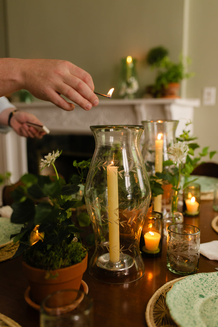 Person lighting a candle in a glass lantern on a table with plants and candles.