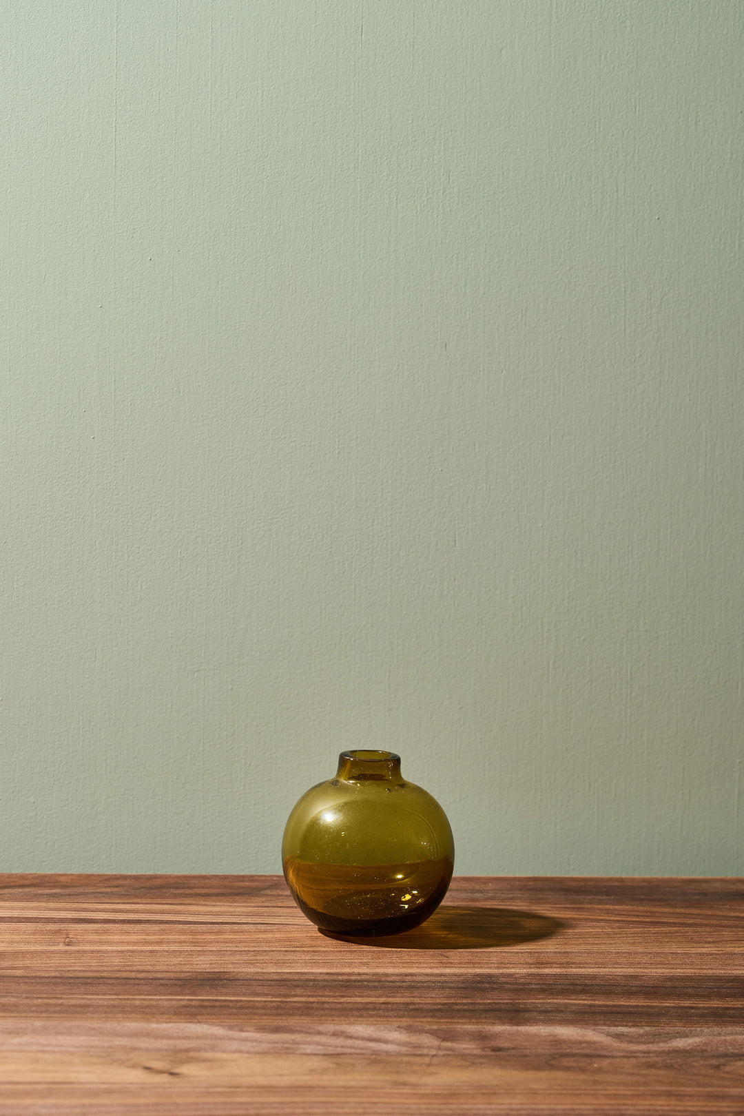 A small round olive green glass vase with a short neck sits on a wooden tabletop against a pale sage green wall.
