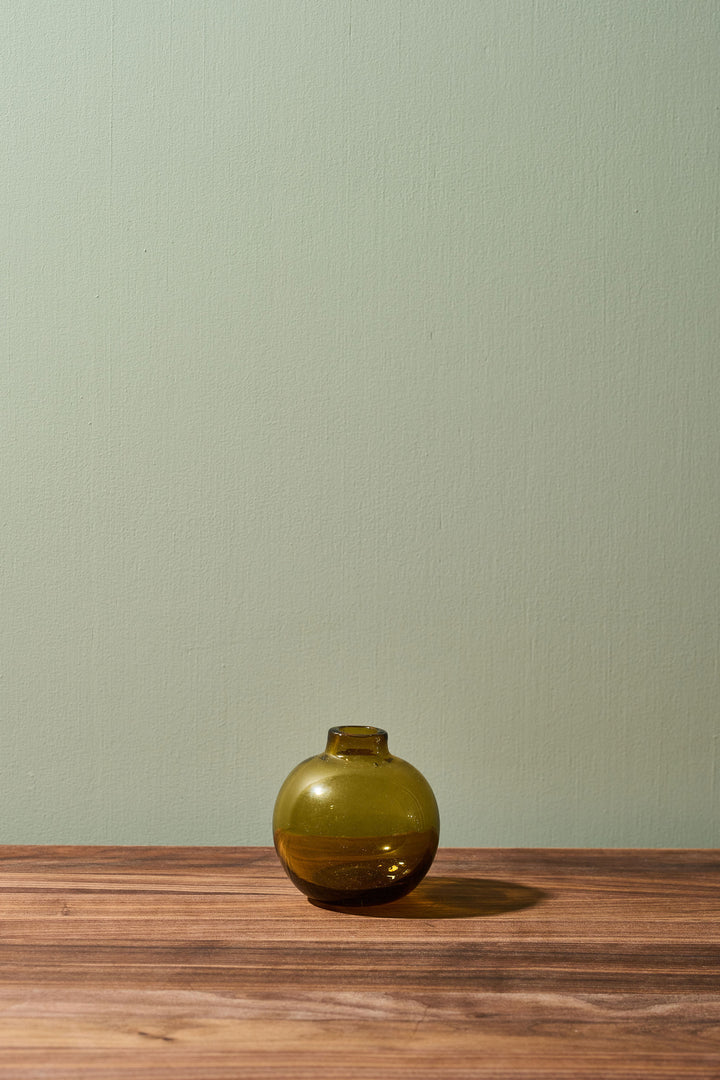 A small round olive green glass vase with a short neck sits on a wooden tabletop against a pale sage green wall.