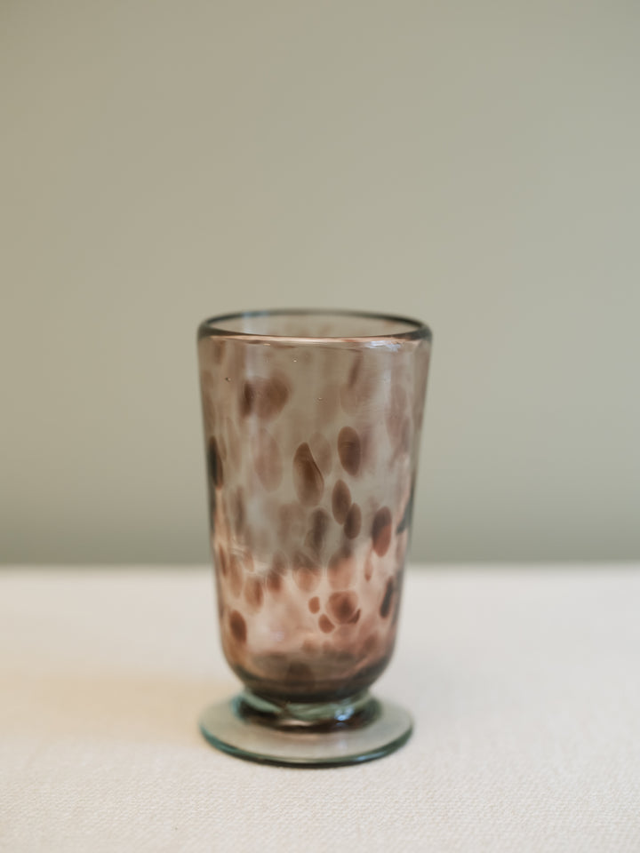 A small clear glass tumbler with a rounded base, featuring a mottled pattern of brown and translucent smoky tones, set against a neutral beige background.