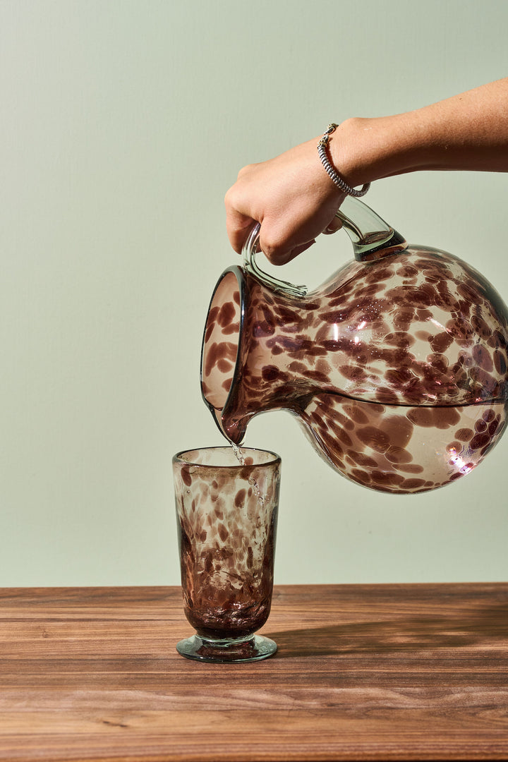 A hand pours liquid from a clear glass pitcher with brown speckled marbling into a matching brown speckled glass tumbler, set on a wooden table against a pale green background.