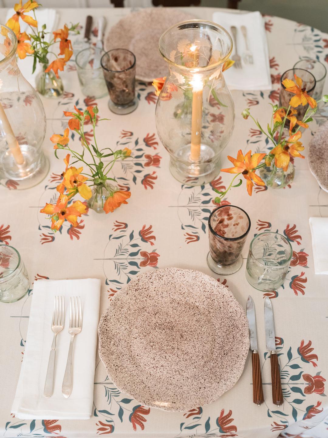 An overhead view of a styled dining table setting featuring a speckled beige plate, silver forks and knife with brown handles, clear glassware, and orange flowers on a floral tablecloth.