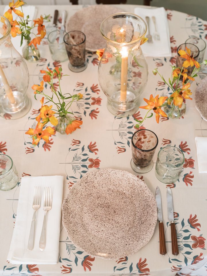 An overhead view of a styled dining table setting featuring a speckled beige plate, silver forks and knife with brown handles, clear glassware, and orange flowers on a floral tablecloth.
