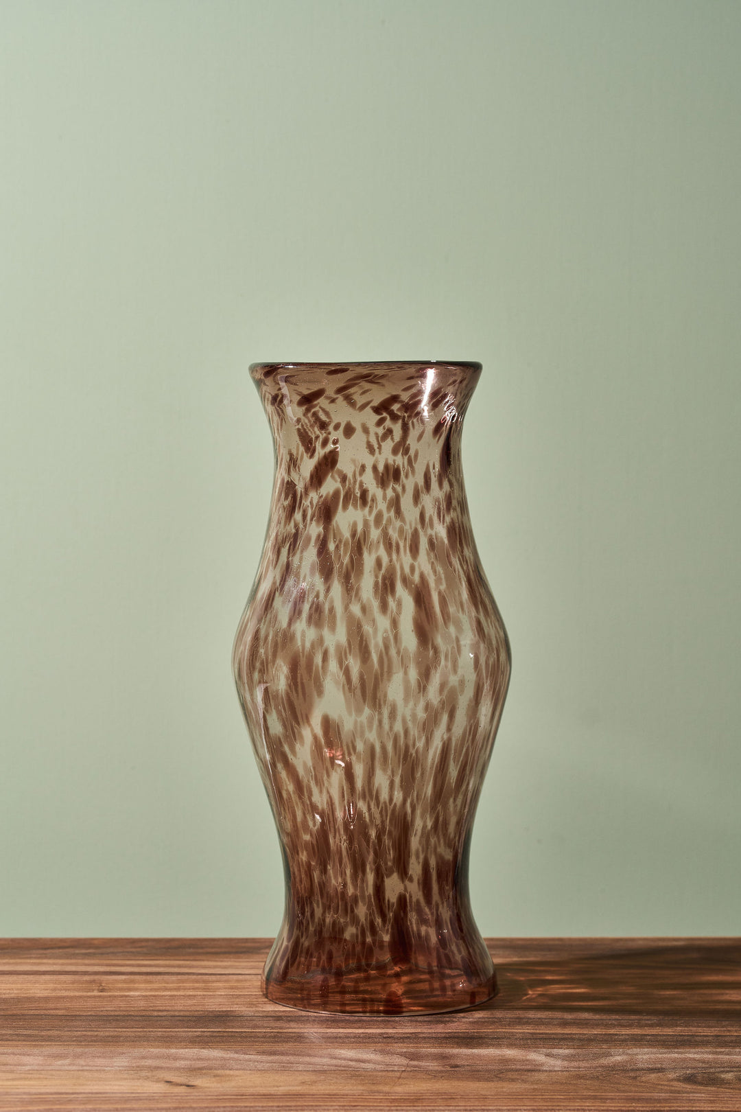 A brown and beige mottled glass vase with a flared rim, standing on a wooden surface against a pale green background.