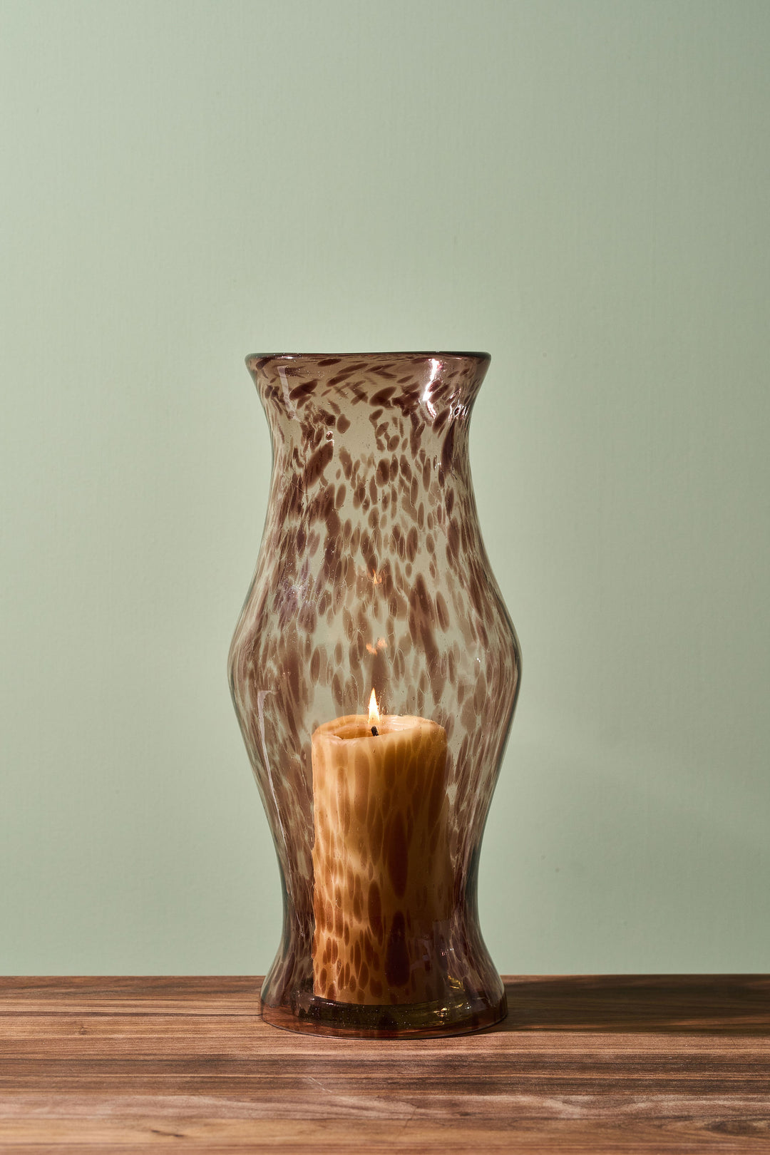 A brown and beige leopard-print glass candle holder vase with a lit pillar candle inside, set on a wooden surface against a pale sage green background.