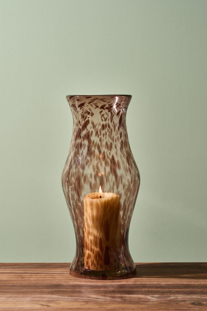 A brown and beige leopard-print glass candle holder vase with a lit pillar candle inside, set on a wooden surface against a pale sage green background.