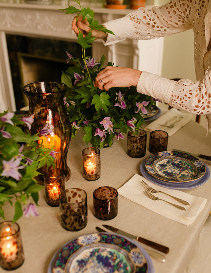 A styled dining table scene with a person arranging greenery and purple flowers in a centerpiece. The table is set with patterned blue and white plates, silverware, amber glass candle holders with lit candles, and a tall amber vase.