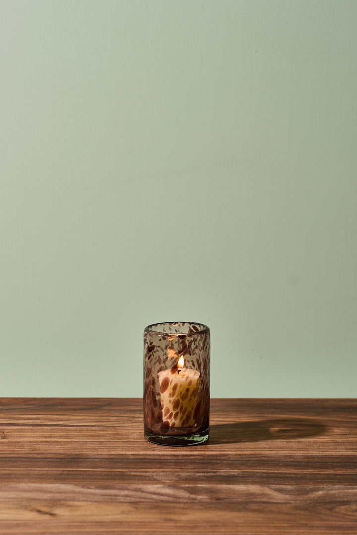 A lit candle in a small textured amber glass holder, placed on a wooden tabletop against a pale sage green wall.