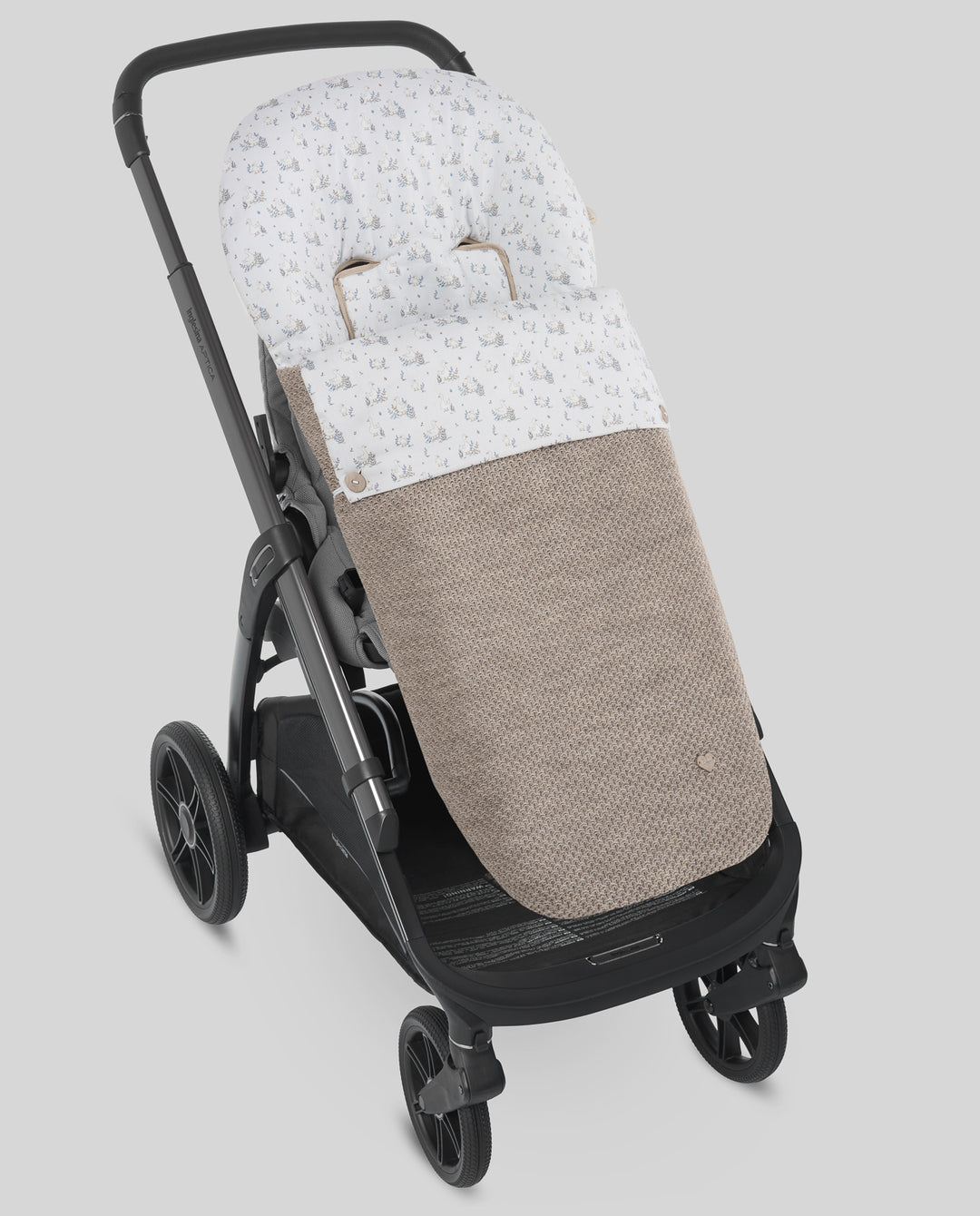 A stroller footmuff with a small floral pattern in white and beige, attached to a black stroller against a solid gray background.