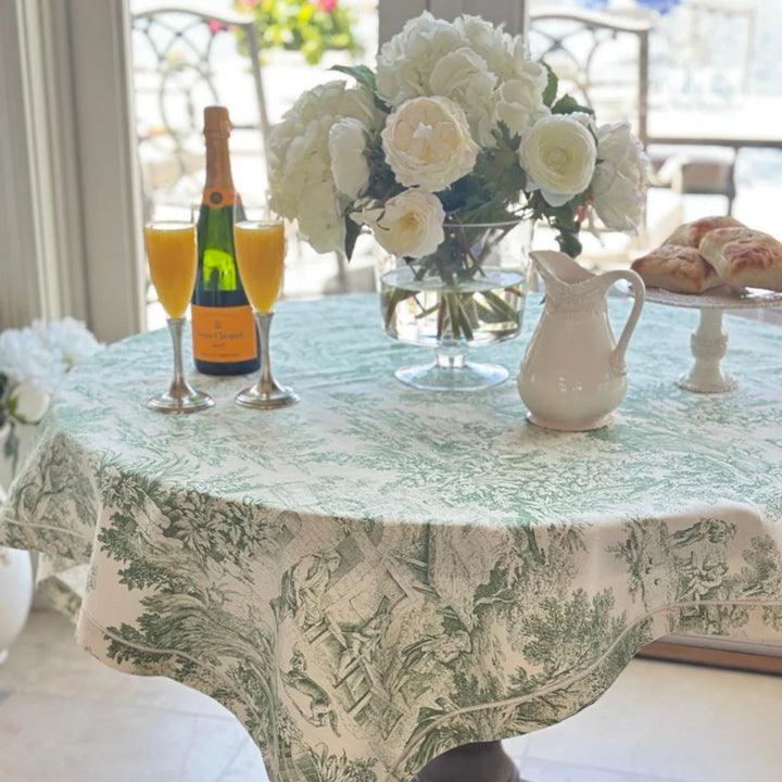 Decorative tablecloth with floral pattern on a table set with a bottle, glasses, and flowers.