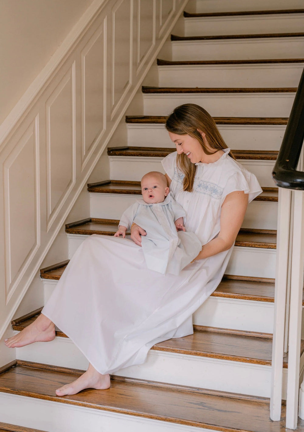 Woman in a white dress sitting on a staircase holding a baby in a matching outfit.