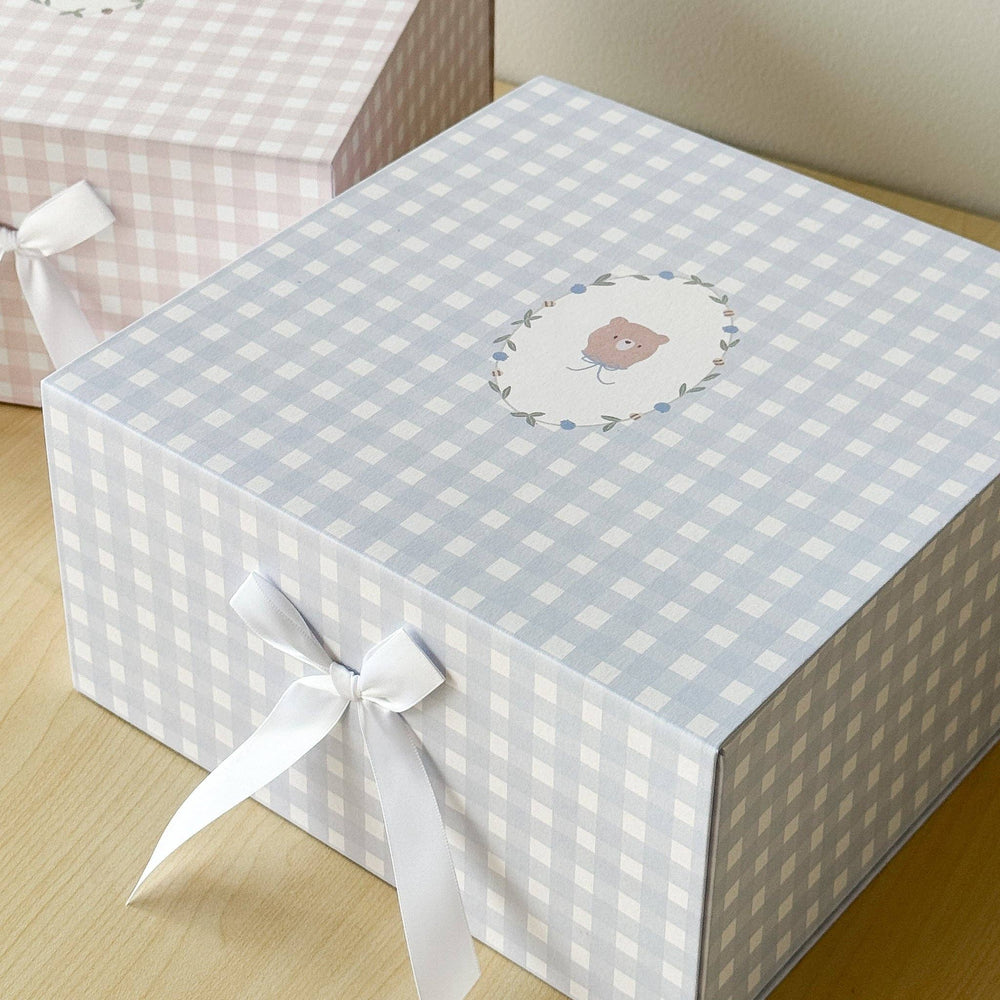 A square gift box with a light blue and white gingham pattern, tied with a white ribbon bow on the front. The top features a small oval illustration of a teddy bear inside a floral border.