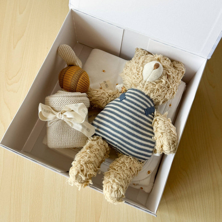 Knitted teddy bear and baby rattle in a white box on a wooden surface