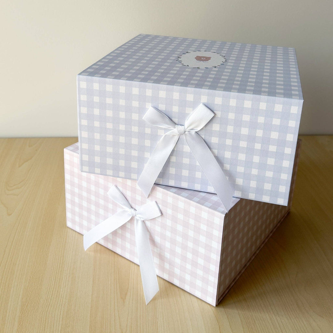 Two stacked gift boxes with gingham patterns, one in light blue and white and one in light pink and white, both tied with white satin ribbons, on a light wood surface against a beige wall.