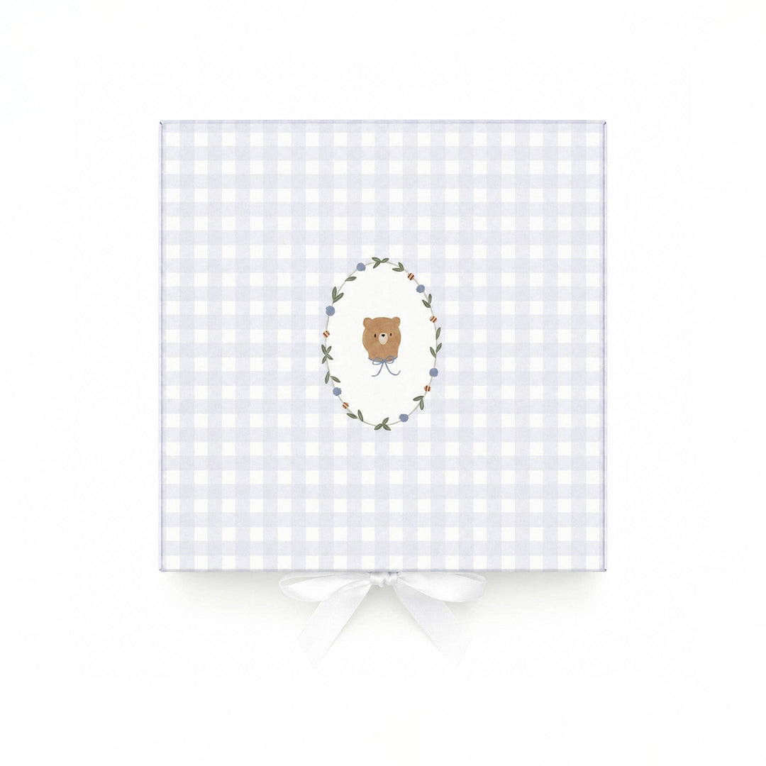 A square gift box wrapped in a light lavender and white gingham pattern, featuring a central illustration of a brown teddy bear inside a floral wreath. The box is tied with a white satin ribbon at the bottom and is photographed against a clean white background.