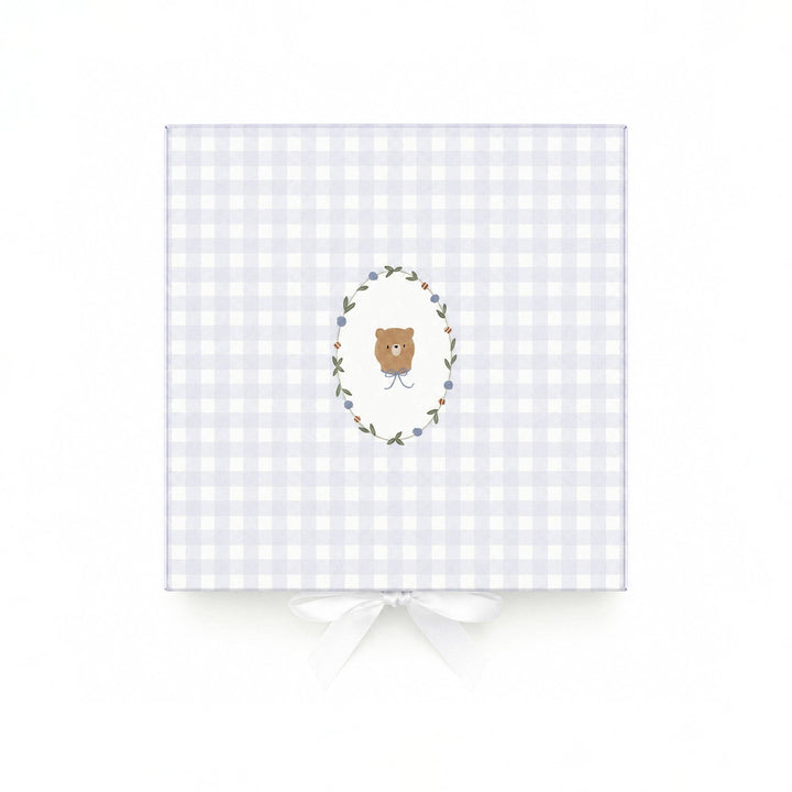 A square gift box wrapped in a light lavender and white gingham pattern, featuring a central illustration of a brown teddy bear inside a floral wreath. The box is tied with a white satin ribbon at the bottom and is photographed against a clean white background.