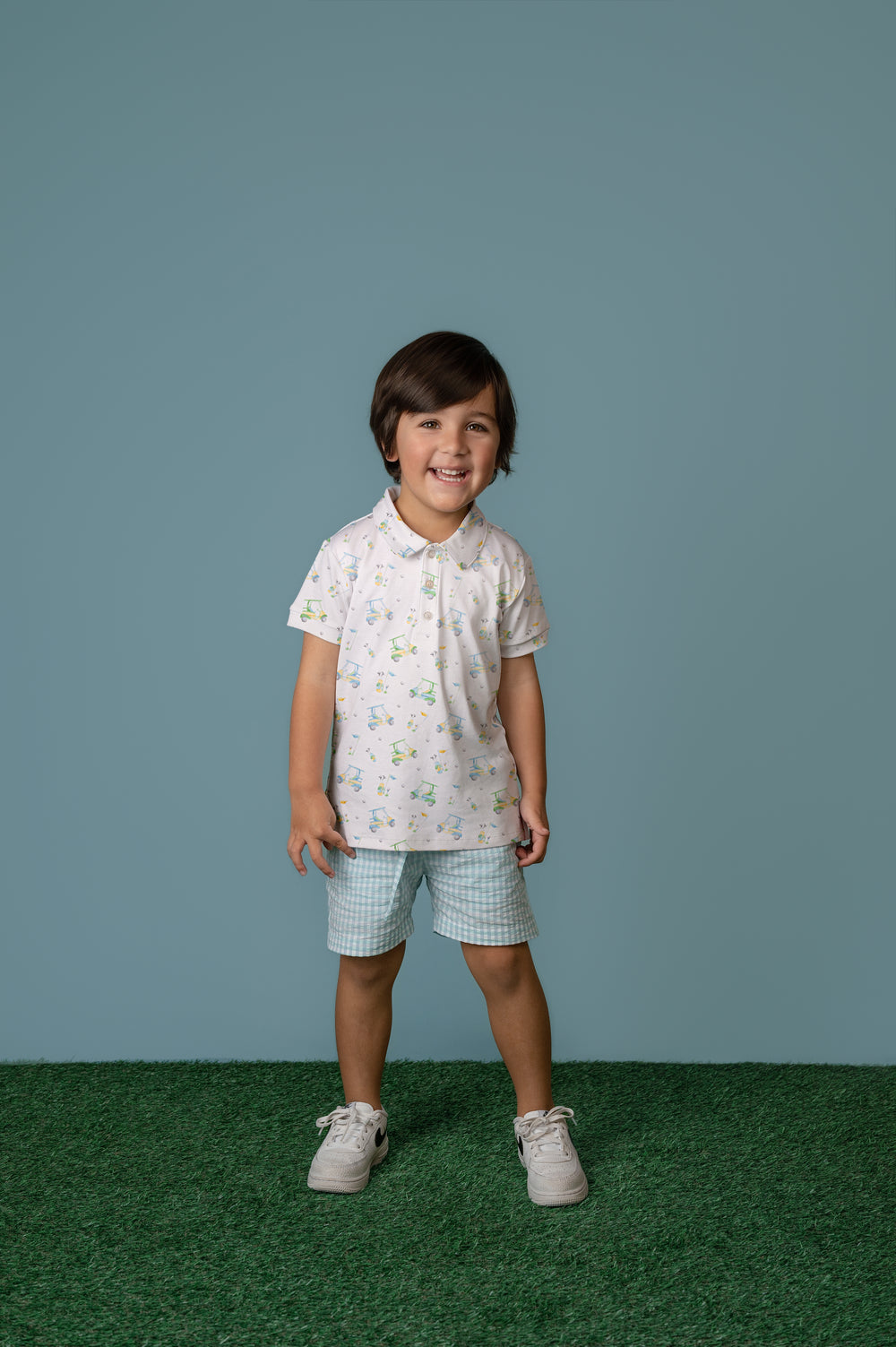 Child wearing a light-colored shirt with a pattern and checkered shorts on a plain background