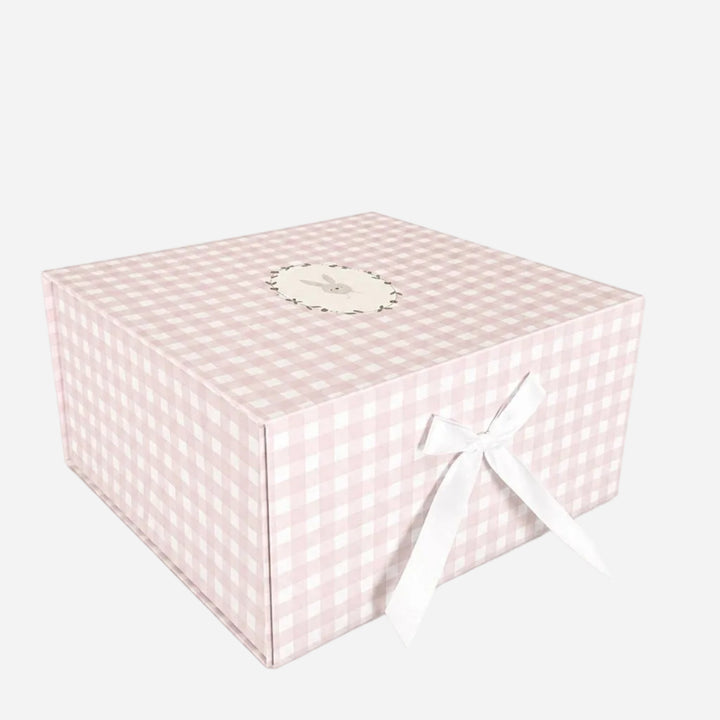 A square gift box with a light pink and white gingham checkered pattern, decorated with a white ribbon bow on the front and a small round label on top, set against a plain white background.