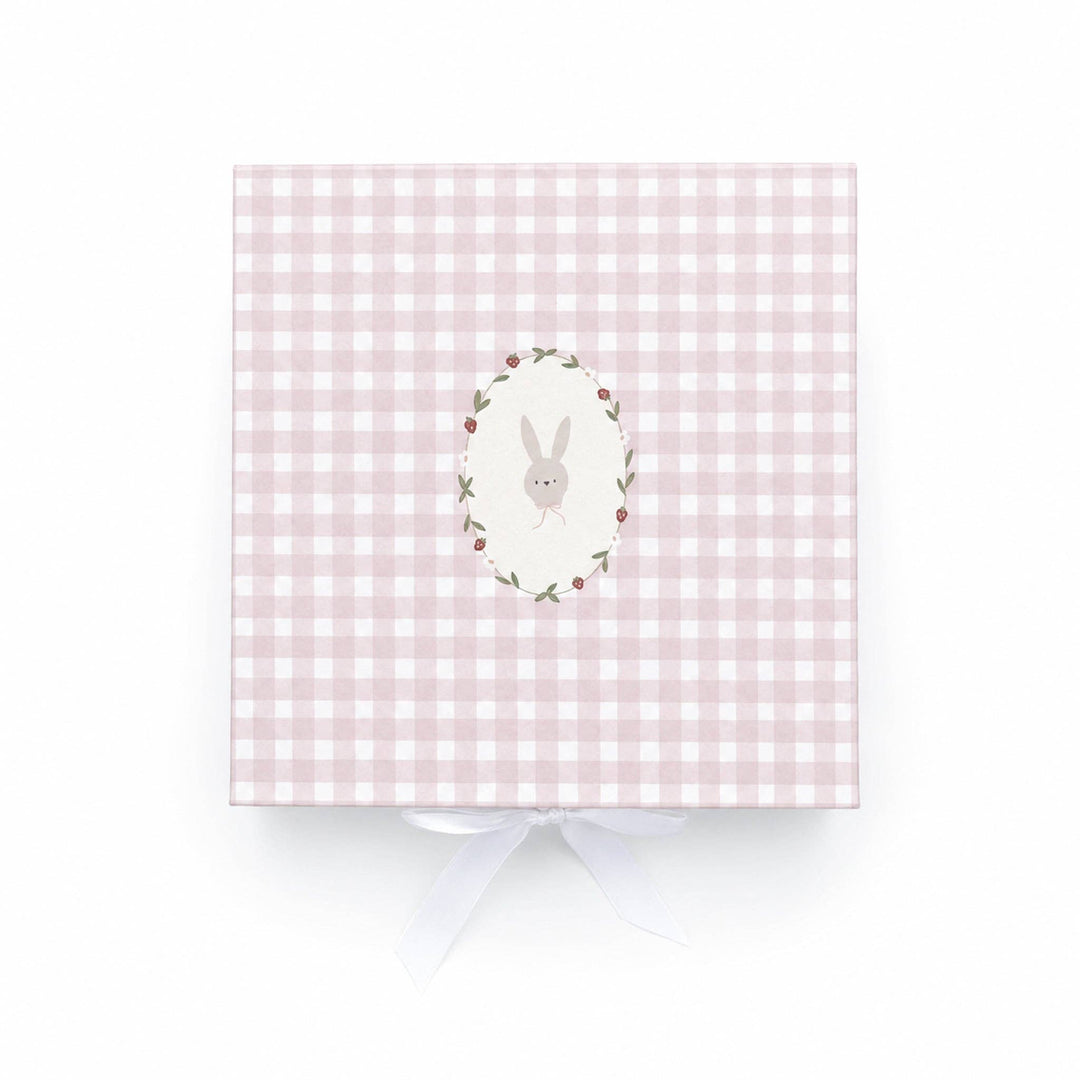 A white plush bunny toy with a pink bow sits inside an open pink-and-white checkered gift box on a light wood surface. A wooden ring and a red strawberry-shaped teething toy are also visible in the box.
