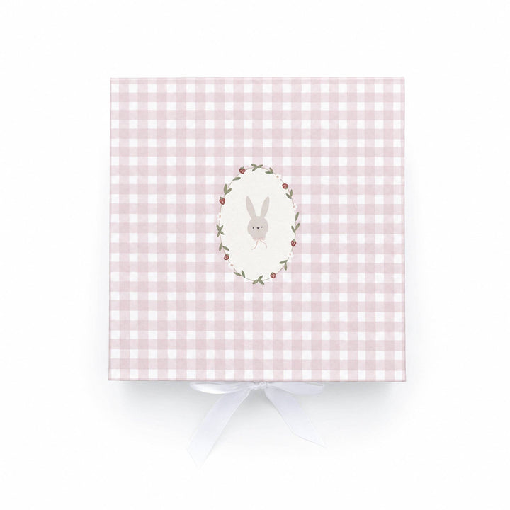 A white plush bunny toy with a pink bow sits inside an open pink-and-white checkered gift box on a light wood surface. A wooden ring and a red strawberry-shaped teething toy are also visible in the box.