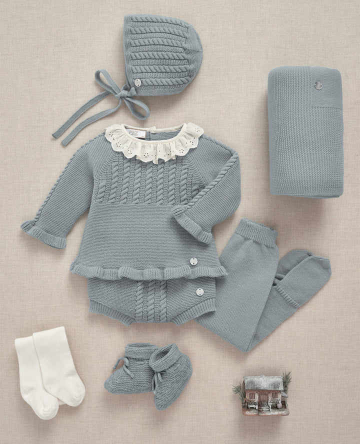 A baby knit outfit set laid flat on a light beige textured background, including a long-sleeve cable-knit romper with a white lace collar, matching hat, booties, socks, and a folded blanket.