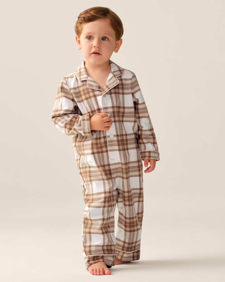 A toddler stands barefoot against a plain light beige studio background wearing a long-sleeve plaid pajama set in white and brown. The pajama top has a collar and front buttons, and the matching pants are shown fully on-body.