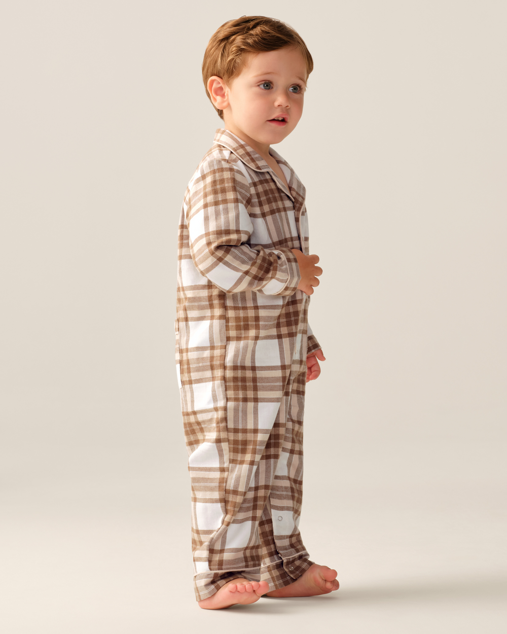 A toddler boy standing in profile wearing a brown and white plaid pajama set against a plain light beige studio background.