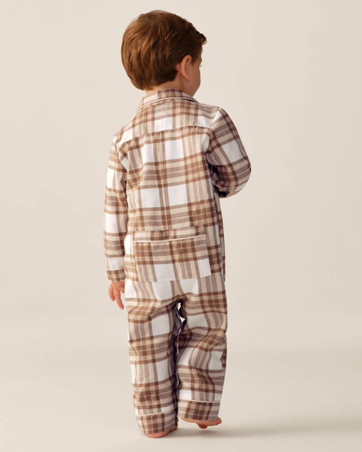 A toddler boy seen from behind wearing a brown, white, and beige plaid long-sleeve jumpsuit, standing against a solid light beige background.