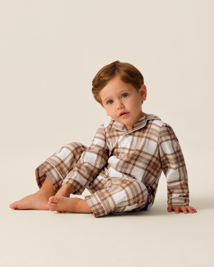 A toddler sitting on a light beige studio floor wearing a brown and white plaid long-sleeve pajama set with matching pants.