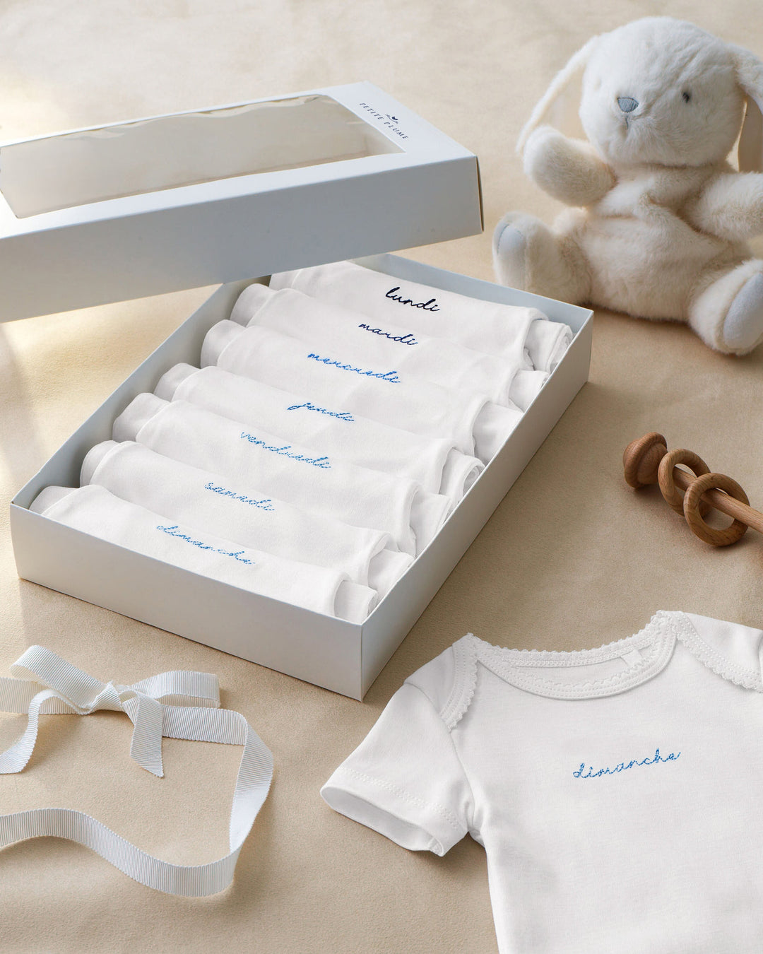 A gift set of white baby clothing and accessories arranged in an open white box on a beige surface, with a plush bunny toy and wooden teething rings nearby. The set includes folded baby socks or mittens with pink script labels, a white bodysuit with pink script text, and a white ribbon bow.