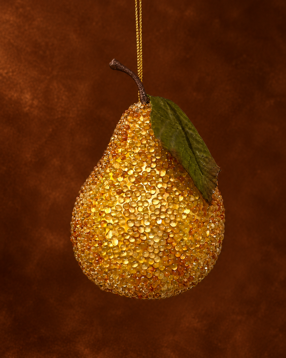 A pear-shaped hanging ornament covered in shimmering gold glitter, with a dark brown stem and a single green leaf, suspended against a warm brown textured background.