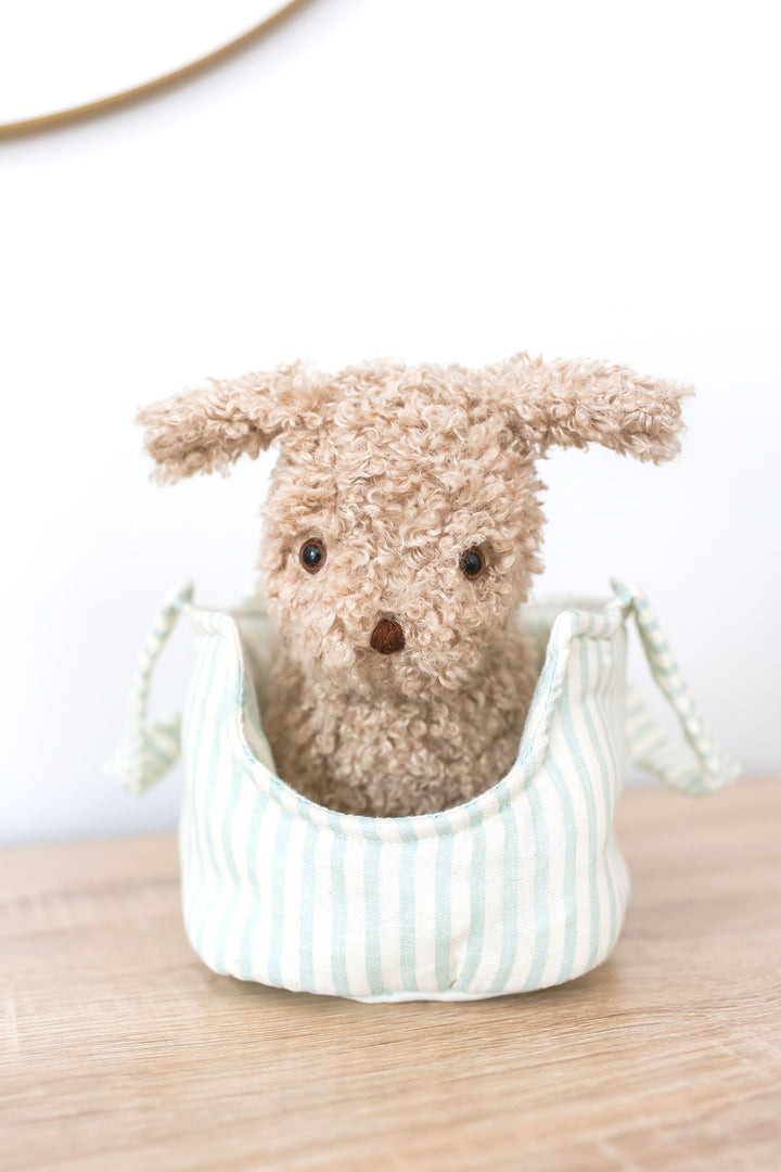 Small plush dog toy in a striped fabric basket on a wooden surface with a white background