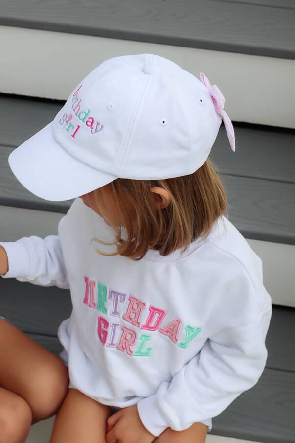 Child wearing a 'Birthday Girl' sweatshirt and cap with a pink bow.