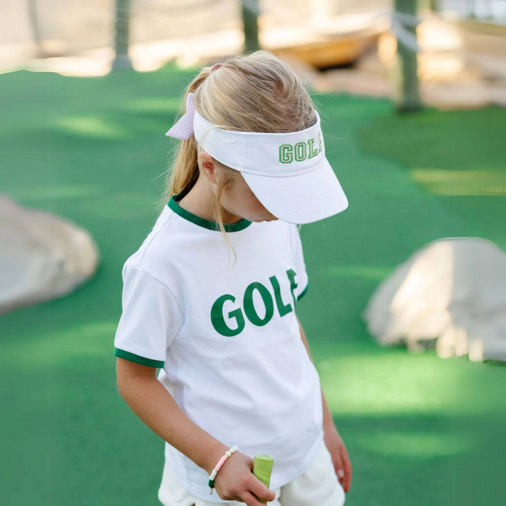 Child wearing a white visor and shirt with 'GOLF' on a green outdoor setting