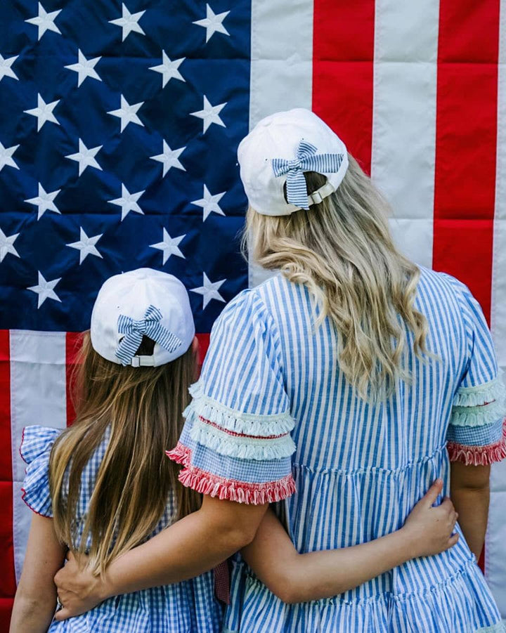 Two women in matching blue and white striped dresses with ruffled sleeves hugging in front of an American flag.