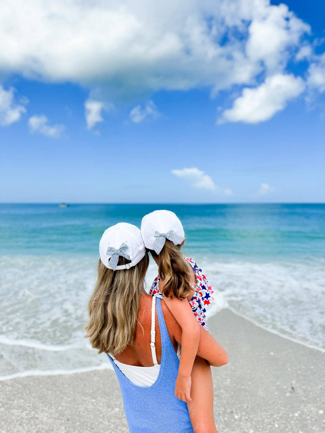 Two women on a beach wearing matching white hats and blue swimsuits, looking out at the ocean.