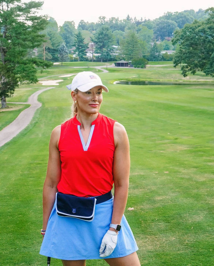 Woman on a golf course wearing a red top and blue skirt with a white cap.