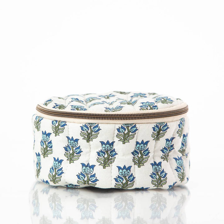 A round quilted cosmetic pouch with a blue and green floral block print on a white background, featuring a brown zipper closure, shown on a reflective white surface.