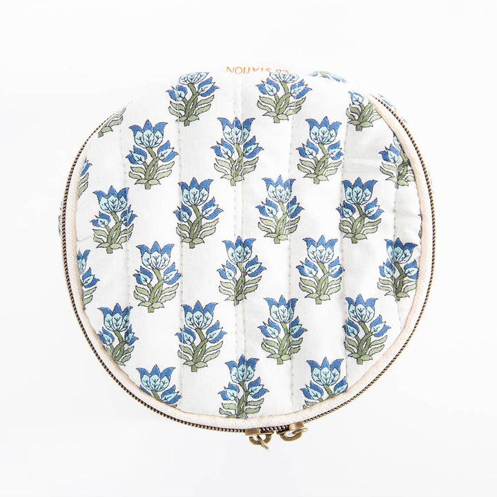 A round quilted pouch with a white background and a repeating blue and green floral block print, finished with a gold-toned zipper.