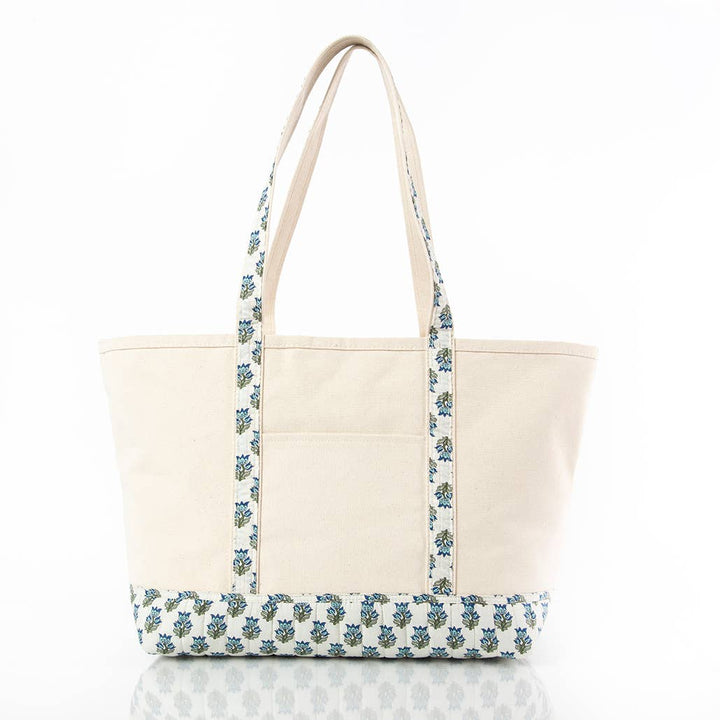 A cream-colored canvas tote bag with blue and green floral block-printed patterned straps and bottom trim, shown on a white background with a soft reflection.