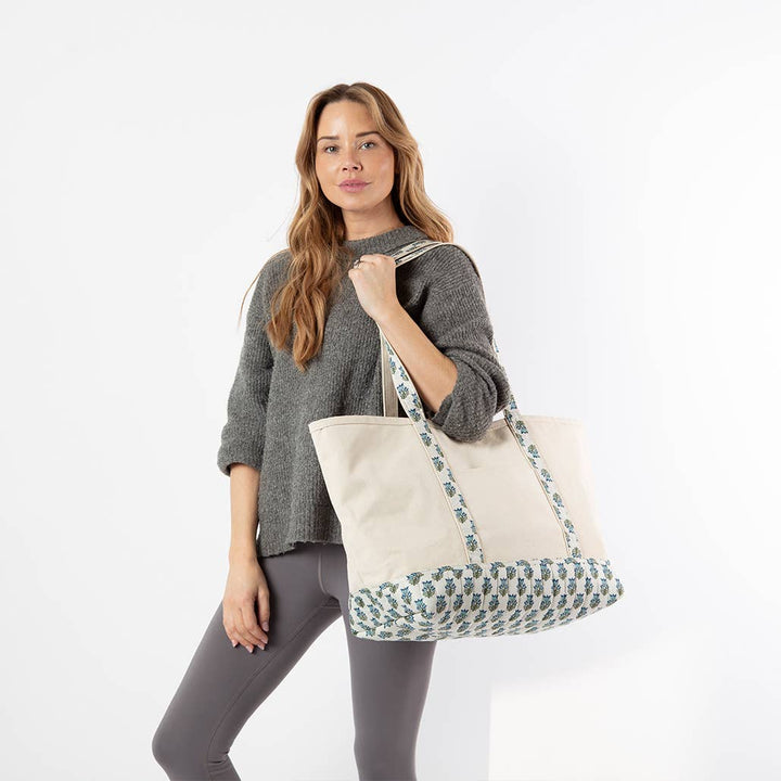 A woman wearing a gray knit sweater and leggings carries a large cream canvas tote bag with a blue and green geometric pattern on the handles and bottom trim, set against a plain white studio background.