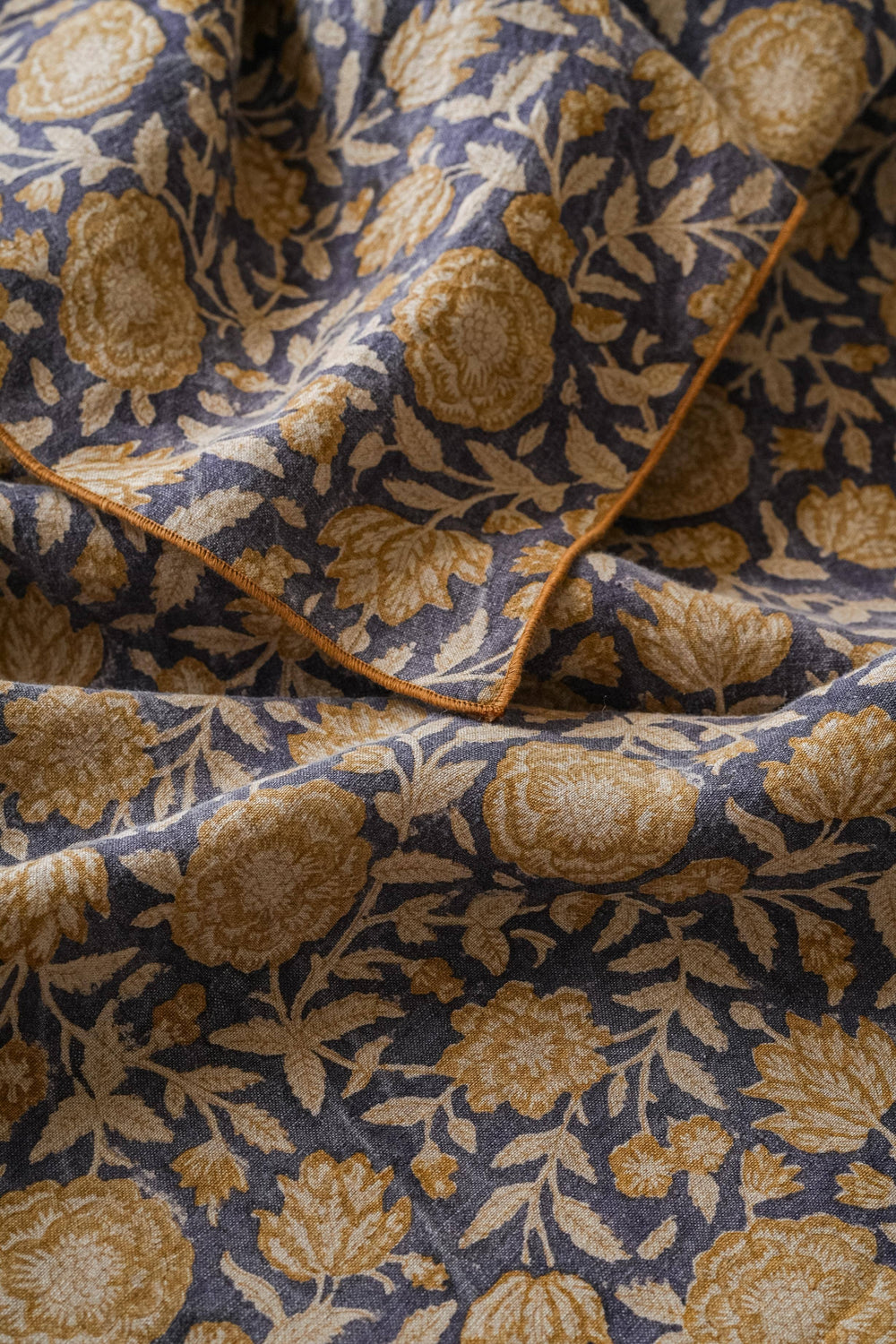 A close-up of draped fabric with a floral pattern in warm beige and muted blue-gray tones, showing the texture and edge stitching.